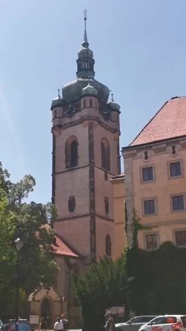 The video shows a church tower with a clock and a spire. There are trees and buildings surrounding the tower. There are a few people visible in the lower part of the frame, and cars are parked in front of the building. The sky is clear and blue.