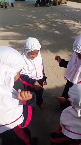 Four young girls wearing white hijabs with red accents stand in a circle on a sandy patch of ground. They are also wearing matching tracksuits with black pants and white and red jackets. One girl is facing the camera, while the others are facing away from the camera. The girls appear to be playing a game. In the background, a motorcycle is parked near a building. Potted plants and landscaping are visible in the background as well. The scene is outdoors, bathed in sunlight.