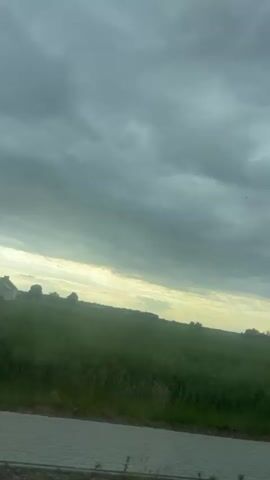 The video shows a landscape view from a moving vehicle. A green field stretches towards the horizon under a cloudy sky. An electrical pole passes by briefly. A paved edge of a road is visible in the foreground.