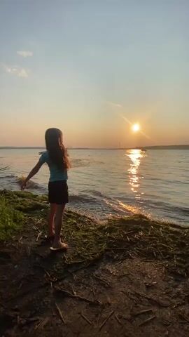 A young girl with long brown hair stands on the shore of a body of water, facing the setting sun. She is wearing a blue t-shirt and a dark skirt. The sun is low in the sky, casting a warm glow over the scene and reflecting on the water. The shore is covered in green moss and debris. A tree with green leaves is visible on the right side of the frame. The sky is clear with a few faint clouds.