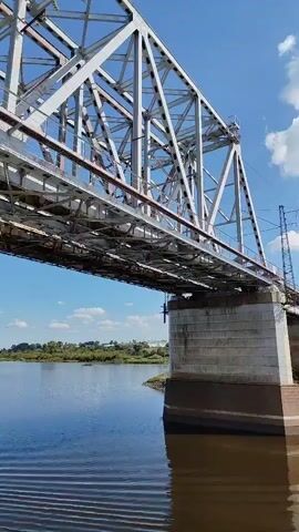 A low-angle, wide shot shows a large steel railway bridge spanning a wide, calm river under a bright, partly cloudy sky. The camera perspective is from a boat or watercraft moving slowly under the bridge, indicated by the slight rippling of the water and the movement of the bridge's structure across the frame. The bridge's complex lattice of steel beams and supports dominates the upper portion of the image, with some overhead wires visible. The concrete piers supporting the bridge are visible in the mid-ground, partially submerged in the brown water. The far bank of the river is lined with green trees and vegetation, with a few buildings and a power line tower visible in the distance. The video is a static shot with no discernible action other than the implied movement of the camera.