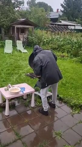 The video shows a person in a black hooded jacket and grey pants standing in a backyard. The ground is wet, suggesting it has recently rained. The person is facing a black grill with the lid open and appears to be placing food onto the grill. To the left of the grill, there is a small pink plastic table with a bowl and some utensils on it. In the background, there is a small wooden shed, two lounge chairs, and a greenhouse. Trees and other greenery surround the yard.