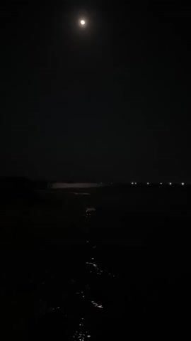 This media file depicts a nighttime scene at Legonipah, Indonesia, with a full moon illuminating the sky. The foreground is a dark expanse of what appears to be wet sand or shallow water, reflecting the moonlight with shimmering highlights. In the middle ground, there's a body of water, likely the sea, with a faint, sandy shore visible on the opposite side. The distant horizon is marked by a line of small, bright lights, possibly from buildings or boats, stretching across the frame. The overall mood is serene and peaceful, with the darkness of the night contrasted by the bright moon and distant lights. There is no visible text in the media file. The time of day is clearly night, indicated by the darkness and the presence of the moon. The weather appears clear, allowing for visibility of the moon and stars. The location cues suggest a coastal area.