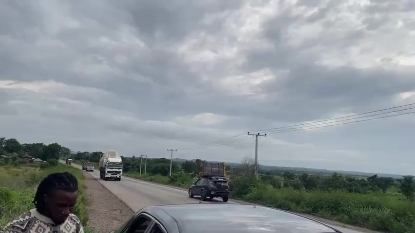 A truck travels down a road under a cloudy sky. A person is partially visible in the foreground, looking down. The truck is white with a black cargo bed, and the cargo is covered with a gray tarp. There are other vehicles on the road and trees on either side. There are also power lines and utility poles visible.