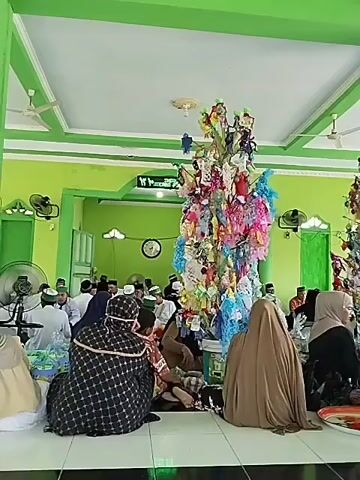 The video shows a gathering of people, primarily women and children, sitting on the floor inside a building with green walls and ceiling. In the center of the room, there is a tall, elaborately decorated structure resembling a tree, adorned with colorful fabrics, ribbons, and small items, possibly ornaments or offerings. Several people are seated around this central structure. Other individuals are visible in the background, also seated, and some appear to be men. The overall impression is of a community event or religious gathering. The lighting is bright, suggesting it is daytime. The floor is tiled with a black and white checkered pattern. Some individuals are wearing traditional Islamic clothing, including headscarves. The atmosphere appears to be calm and communal.