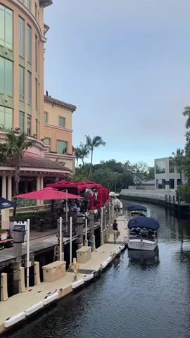 A man stands on a dock next to a covered boat. The boat is in a canal next to a building with red umbrellas outside.