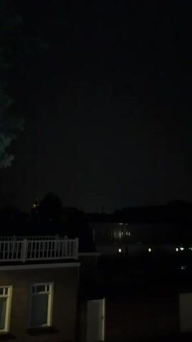 The video shows a nighttime scene with a dark sky. In the foreground, the top of a brick building with white railing is visible. Further in the distance, there are silhouetted buildings and trees. Lights can be seen reflecting on what appears to be water. The overall impression is of a quiet, dark evening.