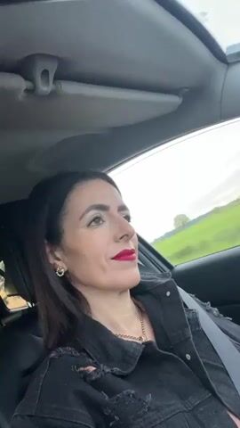 A woman wearing a black denim jacket, a gold chain, and red lipstick is driving a car with a sunroof. She looks relaxed and occasionally looks up at the sky through the sunroof. Music is playing in the background. The car appears to be moving, with glimpses of roadside scenery visible through the windows. The lighting is bright, suggesting it is daytime. The camera is positioned inside the car.