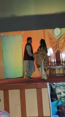 Two individuals are dancing on a stage that is illuminated with rapidly changing colored lights. The woman is wearing a tan floral outfit with gold shoes and the man is in a gray shalwar kameez with a vest. The stage has a green curtain on the left, and a decoration resembling a butterfly with a clock on the right. The stage backdrop is a light blue. A speaker or decorations are visible in the foreground.