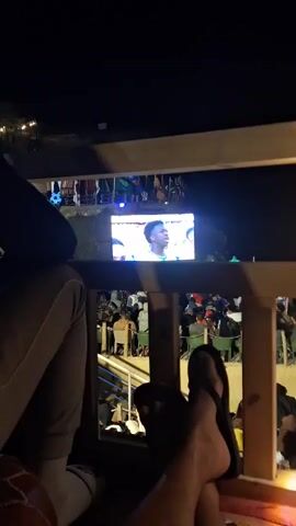 The video is a nighttime shot from a slightly elevated perspective, looking down and across at a large outdoor screen displaying a broadcast. In the foreground, the legs of two people are visible, one wearing flip-flops and the other wearing pants. They appear to be seated, possibly on a balcony or elevated platform, with a railing in front of them. The screen shows people in a crowd, possibly at an event or stadium, with athletes or performers on stage or the field. The lighting is dim, with the screen being the brightest element. Flags of various nations are visible behind the screen, suggesting an international event. The overall atmosphere is casual and social, with people gathered to watch the broadcast.
