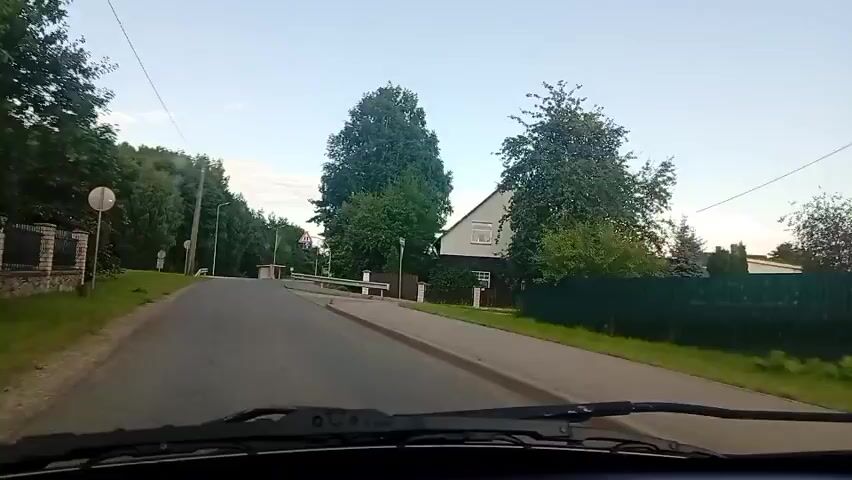 The video captures a view from inside a moving vehicle, likely a car, driving down a paved road. The time of day appears to be late afternoon or early evening, with the sun casting soft light. Lush green trees line both sides of the road. On the right side of the road, a multi-story house with white siding and a dark roof is visible. Several traffic signs are present. One sign, a triangular warning sign with a red border and a white interior, depicts two diverging lines, indicating a road split or junction. Another sign, a blue rectangular sign with a white border, shows a white arrow pointing upwards and to the right with a red diagonal line through it, signifying the end of a route or a mandatory direction. Further down the road, a pedestrian crossing is indicated by white markings on the asphalt. The road itself has a painted white line separating lanes and a white barrier along the right side. Electrical poles and wires are visible along the roadside. The sky is a clear, pale blue with a few wispy clouds. The video is a continuous shot with no discernible audio.
