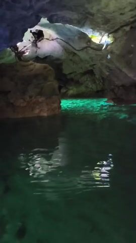 The video shows an underwater cave with glowing green water. A diver with a snorkel and mask is visible in the water, along with some fish. The cave walls are rocky and uneven, and there are lights illuminating the scene.