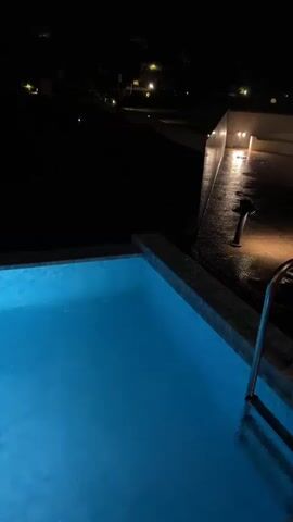 The video shows a nighttime view of two swimming pools. The closest pool is smaller and lit with blue light from below. The camera pans over to reveal a longer pool, in which there are two people swimming. The area around the pool is dark, but there are a few lights in the background illuminating some trees.