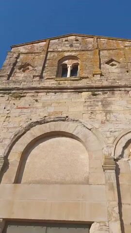 The video shows the facade of an old stone building, likely a church, in Peccioli, Italy. The architecture appears to be ancient, with weathered stone blocks and arched doorways. The sunlight casts shadows, suggesting it is daytime. The sky is a clear blue, indicating fair weather. The building's stone facade has a rough texture, and patches of moss or lichen are visible, hinting at its age. A small, arched window with a central column is positioned near the top of the facade. Below this window, a large, recessed area is framed by an arch, and beneath that, a pair of large, metal doors with numerous rivets are visible. The overall impression is one of historical significance and enduring structure. No people are visible in the frame.