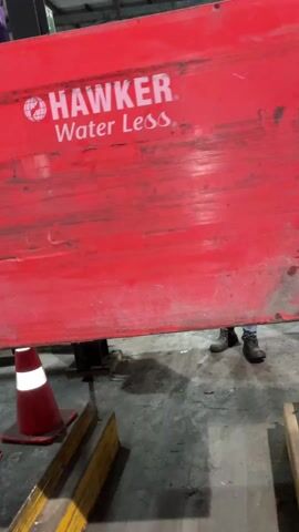 The video shows a forklift operator lifting a large red Hawker Water Less battery using a crane. The battery is slightly scuffed and dirty. It is being lifted from a set of yellow forklift forks sitting on a grey concrete floor. There's also a red and white traffic cone visible. The operator, wearing a safety vest, can be seen in the background. The background reveals the interior of a warehouse.