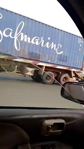 The video, shot from inside a car, shows a yellow van being manually pushed to move. The van is missing its front left tire. The van is being pushed by two men. The van moves and crosses the street. There is a gas station in the background. There is a truck carrying a shipping container parked nearby. Other cars pass.