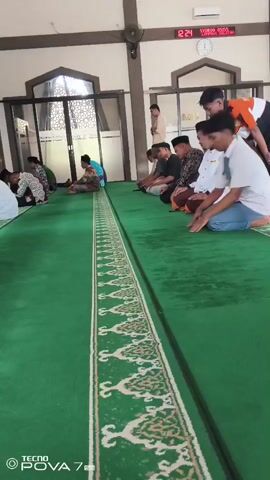 The video shows a group of people, mostly young men and boys, performing Islamic prayer, known as Salah, inside a mosque. The floor is covered with a green carpet with a decorative border. The individuals are arranged in rows, facing the direction of the Kaaba in Mecca. Most of them are in the prostration position (Sujud), with their foreheads touching the ground. The environment appears to be indoors, with a high ceiling and some decorative elements on the walls. There are windows or glass partitions visible, and a digital clock displaying 12:24 is mounted on the wall. One person in the foreground is wearing a white t-shirt and blue jeans, while others are in religious attire such as kufis (skullcaps). The lighting suggests daytime. A person in an orange and white shirt is standing at the back, seemingly observing.