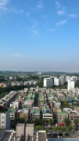 This is a vertical, overhead aerial shot of a dense urban environment under a clear blue sky with scattered white clouds. The foreground is dominated by rows of buildings with predominantly green and orange roofs. These buildings appear to be multi-story residential or mixed-use structures, packed closely together with narrow streets running between them. Cars are visible on these streets. Further in the background, larger, modern white apartment buildings rise above the smaller structures. Beyond the apartment buildings, a green, tree-filled landscape stretches to the horizon, with some distant buildings visible. The overall impression is a sprawling cityscape with a mix of older and newer architecture. There are no people or animals visible in the frame, and no specific actions or interactions are depicted.