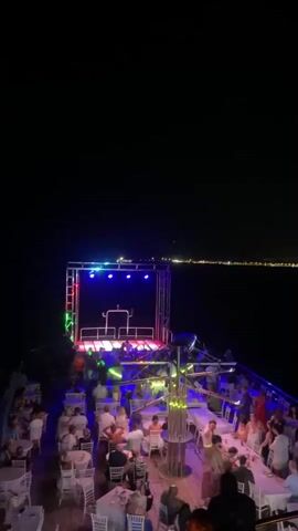 A nighttime aerial view of a party on a boat shows people dancing and dining on a deck illuminated by colored lights. A DJ booth is set up on a raised stage at the front of the boat, with a cityscape visible in the background across a body of water.