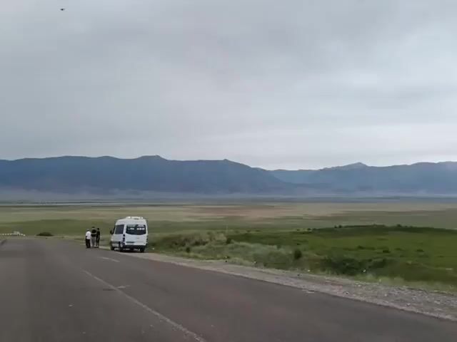 A wide shot captures a desolate landscape under a cloudy sky. A paved road runs diagonally across the foreground, with a white camper van parked on its right shoulder. Three individuals stand near the van. Beyond the road lies a vast expanse of green grassland dotted with small bushes. In the distance, a range of imposing mountains stretches across the horizon, their peaks softened by the atmospheric haze. The overall impression is one of remoteness and stillness.