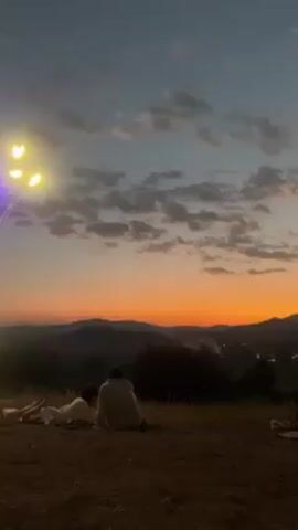 The video shows a group of people gathered on a hilltop at sunset. Silhouettes of the group are visible against the horizon, some sitting on blankets or mats. The sunset paints the sky in hues of orange and red, with clouds scattered across the sky.  In the distance, there are mountains and a valley with lights suggesting a town or city. The foreground is a dirt or grassy area.