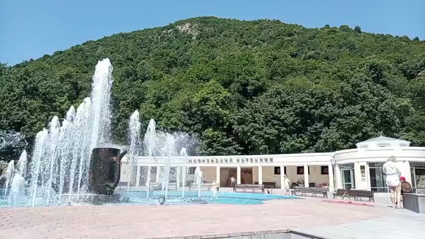 The video shows a wide shot of a public park with a large fountain in the foreground. The fountain has multiple jets of water spraying upwards, and a dark, sculpted object resembling a jug is placed in the center of the main jet. Behind the fountain, there is a white building with a row of arched openings and benches in front of it. The building has text above the arches, which appears to be in a Cyrillic script. A forested hill rises steeply behind the building. Several people are visible: one person in a white top and shorts stands near the building, facing away from the camera with arms raised. Another person in darker clothing stands to the right of the building, partially obscured. Two people are walking near the fountain in the mid-ground, and a few more figures are visible under the arches of the building. The ground in the foreground is paved with light-colored bricks. The sky is clear and blue. The overall impression is a sunny day in a well-maintained park area.
