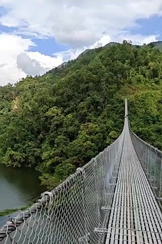 This is a first-person perspective video taken from a suspension bridge overlooking a large body of water surrounded by lush green hills. The bridge is made of metal and has a mesh railing. The water is dark and calm with some patches of green aquatic plants. In the distance, there are mountains covered in dense forest. The sky is partly cloudy with white clouds against a blue background. There is a single person visible on the bridge in the far distance. There is also a small boat or raft in the water. The video pans slowly to the left, revealing more of the lake and the surrounding landscape. The overall impression is one of natural beauty and tranquility.