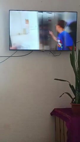 A television hangs on a white wall, with various cords connected to it. The TV screen displays a scene of two boys in an elevator. One boy is wearing a blue shirt, and the other is wearing a yellow shirt and orange shorts; both are carrying white shopping bags. Below the TV, there is a wooden table draped with a dark purple cloth. A potted snake plant stands on the table. In the foreground, a person with dark hair and a beard is lying down, looking at the TV.