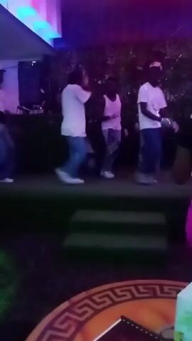 Here is a description of the video:

The video appears to be taken at an outdoor event at night, with colorful lighting creating a party atmosphere. Five people are on a small stage with a faux grass covering. They are dressed casually in white shirts and jeans, and they appear to be dancing in sync. The stage is illuminated with both blue and purple lights. There's a table visible in the foreground.  The table has a decorative circular top with a Greek key pattern. Behind the stage is a wall covered in greenery.