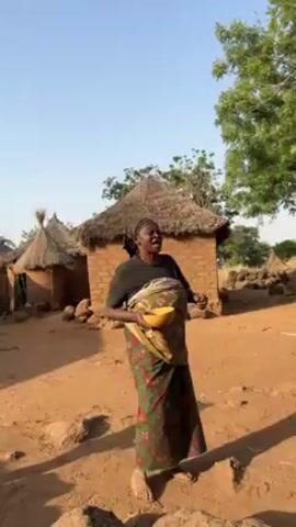 A woman is the focal point of the video, standing outdoors with a bowl held in her arms. Her dark hair is pulled back, and she's wearing a dark top and a colorful patterned skirt. A cloth is wrapped around her upper body, possibly holding something. She appears to be speaking or singing, looking upwards.

The setting is a rural area, evident from the traditional mud huts with thatched roofs in the background. A leafy tree is to her right, and the ground is dry and reddish-brown. The sky is clear and bright, suggesting it's daytime. The video focuses on the woman as she engages in what seems like a performance or address, likely taking place within her village.