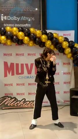 The video shows a male performer dressed as Michael Jackson, wearing a black sequined jacket, black pants with white socks visible at the ankles, and black shoes. He is holding a microphone and singing. He is standing in front of a backdrop with a "MUVIX CINEMAS" logo and a banner that appears to say "Michael." A large arch of black and gold balloons is behind him. To his right, a speaker is visible. Above the balloons, a sign for "Dolby Atmos Sonido Envolvente" can be seen. The performer is in mid-dance pose.