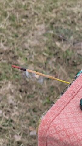 The video shows a lit incense stick with a red tip and a yellow stem, held against a backdrop of dry grass and a pink, textured surface. Smoke is rising from the burning tip of the incense stick. The camera appears to be handheld, with a slight blur in the background.