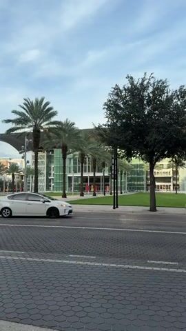 A white car is driving down the street in front of a modern building with large windows and palm trees. The sky is cloudy.