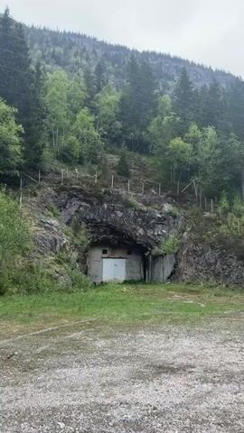 The video shows a historical bunker. The bunker is located in a rocky hillside covered with trees and vegetation. The bunker has a white door and is surrounded by concrete structures. There is a gravel area in front of the bunker. There is a tall fence above the bunker on the hillside.