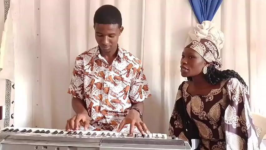 A young man in a patterned short-sleeved shirt is playing a keyboard. A woman wearing a brown patterned dress and headscarf is standing next to him and appears to be singing. They are both in front of a backdrop consisting of white curtains with blue and white decorations. The keyboard is silver and black. The man is focused on playing, while the woman's eyes are directed forward as she sings.