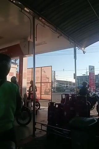 The video shows a gas station with several motorcycles and people. In the foreground, a person on a motorcycle wearing a green jacket with the "Gojek" logo is visible from the back. They are wearing a black helmet. To their left, another person in a black helmet and a green shirt is also on a motorcycle. Further in the background, there are more motorcycles and people at the gas pumps. The gas station has a red and white sign with text on it, including "PERTALITE". There are also large purple gas cylinders visible on the right side of the frame. The environment appears to be outdoors, with a clear sky. The video switches to a close-up of a motorcycle's dashboard and ignition, showing a key inserted. A blue plastic bag is hanging from the motorcycle.