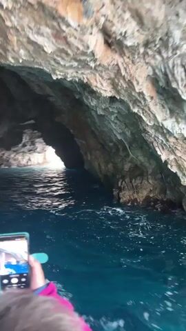 The video shows a person on a boat entering a cave. The boat is moving through blue water, creating white wakes. The cave entrance is visible as a bright opening, framed by the dark, rugged rock formations of the cave walls and ceiling. The rock shows variations in color and texture, appearing weathered and slightly damp. A hand holding a smartphone is visible on the left side of the frame, apparently recording the scene. Other parts of the boat can be seen as well.