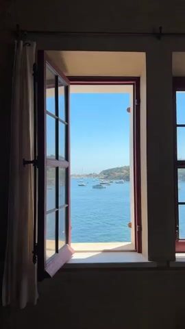 The video shows a view through an open window. The window has a red frame and is looking out onto a body of water, likely the sea. There are several boats, appearing to be sailboats and yachts, anchored in the water. In the background, there is a hillside with vegetation and buildings. The sky is a clear blue. A white curtain is partially visible on the left side of the window. The overall scene is tranquil and suggests a coastal or island location.