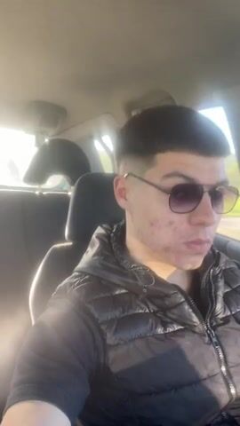 The video shows a young man in the driver's seat of a car. He's wearing sunglasses and a black puffer vest. The car appears to be moving, as scenery is visible through the window. The interior of the car is visible, including the steering wheel, dashboard, and rearview mirror. An air freshener hangs from the rearview mirror. The scenery outside includes green fields and hills.