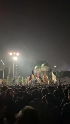 The media file captures a nighttime outdoor scene filled with a large crowd of people. The subjects are primarily young adults, many wearing dark clothing, gathered together. Numerous flags of various colors, some with symbols and text, are being held aloft and waved. In the background, illuminated by bright spotlights, there is a building with distinctive green domes, and further beyond, city lights and structures are visible, suggesting an urban setting. Smoke or mist is present, adding to the atmosphere and diffusing the light.

The activity taking place appears to be a rally, protest, or organized gathering, indicated by the large assembly of people, the presence of flags, and what appears to be a banner with text in the background. The time of day is clearly night, as evidenced by the darkness and artificial lighting. The weather seems to be clear enough for an outdoor event, though the mist might suggest high humidity or the presence of smoke machines.

Notable details include the central focus on the crowd and the flags, the bright stage lighting illuminating the area, and the distinctive architecture of the building with green domes. The mood conveyed is one of collective action and possibly excitement or determination, given the density of the crowd and the active display of flags.

Visible text includes writing on some of the flags and the banner in the background. While most of the text is difficult to read due to the image resolution and angle, some phrases are discernible, such as "PEMBENG" and what might be "SEJARAH" on the banner. The location context suggests this event is taking place in Central Jakarta, Indonesia.