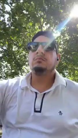 A man wearing a white polo shirt and sunglasses looks into the camera, raising a hand and then giving a thumbs-up gesture. He seems to be moving, possibly on a bicycle or scooter, with trees and the sky visible in the background, along with power lines. The sun is bright, creating a glare. The video appears to be shot in a residential or suburban area.