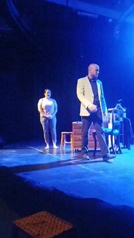 Two actors are on a dark stage illuminated by blue and white spotlights. A man in a light-colored suit jacket and dark pants stands center-right. To his left and further back, a woman in a light-colored t-shirt and grey pants stands facing away from the audience. A wooden dresser with drawers sits between them. The man is looking to his right, and the woman is looking forward. The stage floor is dark blue and reflects the lighting.