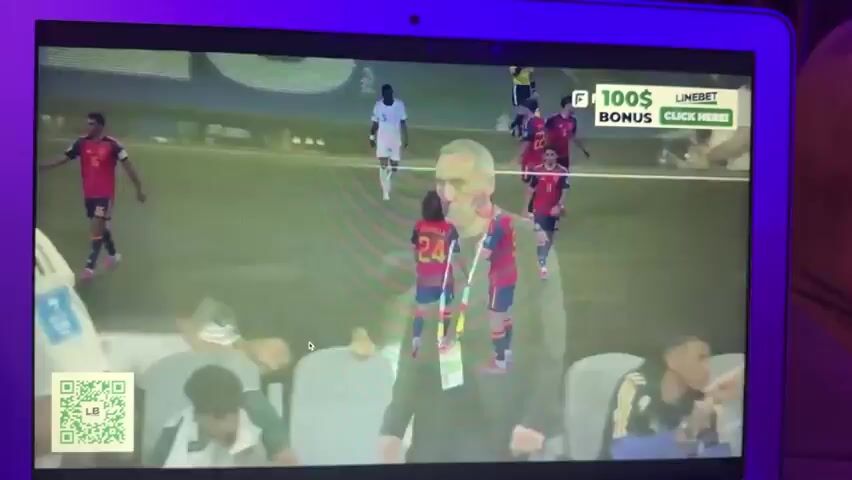 The video shows a laptop screen displaying a soccer match. The match appears to be between Spain and Saudi Arabia. The score is 4-0 in favor of Spain. Several players in red and yellow uniforms are visible on the field, as are a few players in white uniforms. A scoreboard at the bottom of the screen displays the final score and lists the goal scorers. There is also an advertisement banner at the top right of the screen. The scene is dimly lit, and the laptop is positioned on a couch.