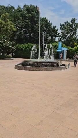 A video pans across a paved courtyard with a tiered fountain in the center. Several streams of water arc upwards from the fountain. Lush green trees and bushes surround the courtyard. A white pole stands behind the fountain. To the right of the fountain, a small blue and white structure is visible, with two people standing near it. A bench is also located to the right of the fountain. A person walks across the paved area to the right side of the frame.