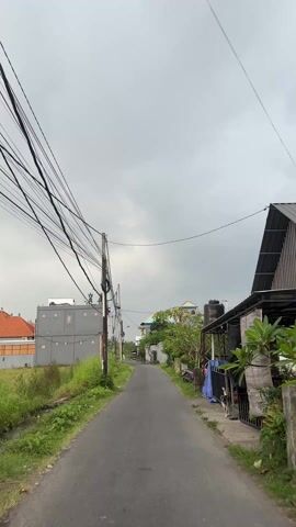 The video shows a first-person perspective of someone riding a motorbike down a narrow paved road lined with houses and vegetation on either side. The sky is overcast and grey. Power lines crisscross above the road, attached to utility poles. The houses are a mix of grey and white, with some traditional Balinese architectural details visible. Some shops appear to be closed, with the shutters down. There are patches of overgrown grass and weeds along the edges of the road. The person riding the motorbike can be seen reflected in the side mirror, and there's another motorbike rider in the distance.