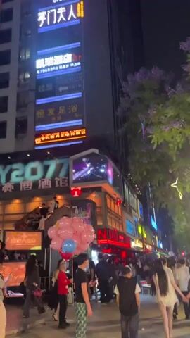 The video shows an outdoor nighttime street scene bustling with people walking on a wide sidewalk. A tall building with multiple signs in Chinese script stands on the left. There are numerous pink and blue balloons tied to something near the foreground. Many shops and establishments line the street, lit with neon lights and other colorful signage. People are of various ages and are dressed in casual clothing. Many seem to be in groups or couples, walking and talking. The overall atmosphere appears lively and festive. There are trees with purple blossoms and decorative lights along the street, adding to the ambiance.