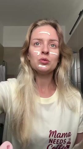 A young woman with blonde hair is applying white cream to her face. The cream is applied in stripes on her forehead, cheeks, and chin. She is holding a tube of cream in her left hand and is using her right hand to spread the cream on her face. She is wearing a light yellow t-shirt with text that says "Mama Needs Coffee". The background appears to be a bathroom with a shower curtain visible.