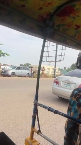 The video is filmed from inside a yellow motorized tricycle, looking forward. The interior of the tricycle is decorated with a floral fabric. Outside the tricycle, there is a paved road with several vehicles including other tricycles and a silver car. In the background, there are buildings, trees, and a billboard. A few people are visible on the side of the road. The weather appears to be overcast.