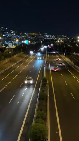 A high-angle drone shot shows a multilane highway at night with traffic. Cars with their headlights on drive in both directions. The road is well-lit by streetlights, and the background shows a city skyline with illuminated buildings. The overall scene is dark with the exception of the artificial lighting from the cars and the city.