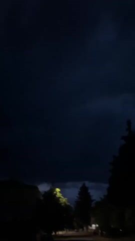 The video shows a view of the sky at night. The sky is dark and cloudy. There are a few trees and buildings visible in the foreground, and a street light is visible, with its light shining through the leaves of a tree.